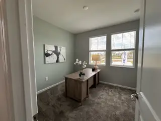 A cozy home office with soft gray walls, a wooden desk, carpeted flooring, and natural light from large windows.
