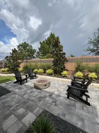 A cozy patio with Adirondack chairs, stone fire pit, and lush landscaping against a brick wall.
