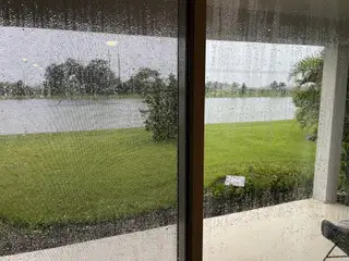 A serene view of a rainy day through sliding glass doors, overlooking a lush green yard by a peaceful waterway.