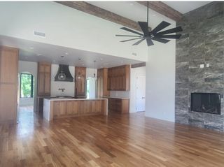 An elegant open space with wooden beams, large fan, wood floors, and a stone fireplace. Modern kitchen with natural light.