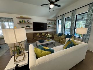 A stylish living space with a white sofa, colorful pillows, a ceiling fan, and a TV mounted on a built-in shelf wall.