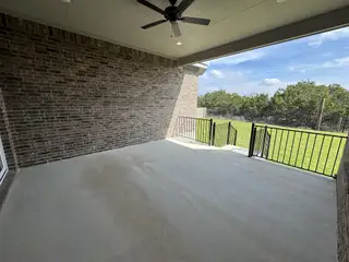 A spacious covered patio with brick walls, ceiling fan, and a view of a lush green yard through black metal railings.