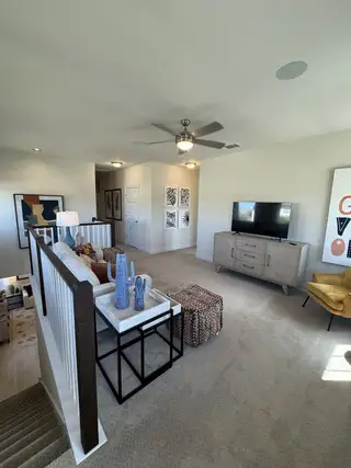 A cozy living space with a modern ceiling fan, plush carpet, and stylish decor, including a TV console and vibrant artwork.