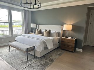 Model Home A serene primary bedroom with a plush upholstered bed, wood accents, and large windows overlooking a scenic backyard.