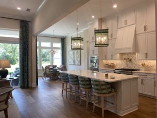 Model Home A spacious kitchen featuring a large island with elegant pendant lighting, white cabinets, and a dining area overlooking greenery.