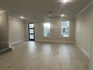 A spacious living room featuring light wood flooring, modern ceiling fan, and abundant natural light from multiple windows.