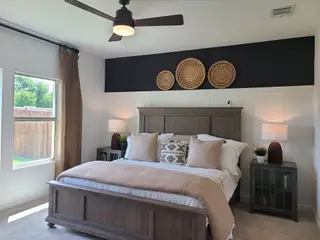 Model Home A cozy bedroom with a wooden bed, earthy tones, decorative wicker accents, and a ceiling fan. Large windows offer a garden view.
