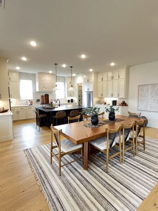 A spacious, modern kitchen with a large wooden dining table, striped rug, pendant lighting, and white cabinetry.