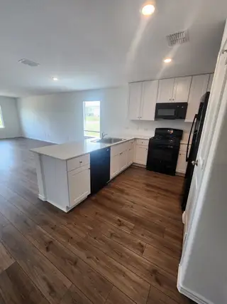 Model Home A modern kitchen with sleek white cabinets, dark appliances, and laminate wood flooring, opening to a bright, spacious living area.