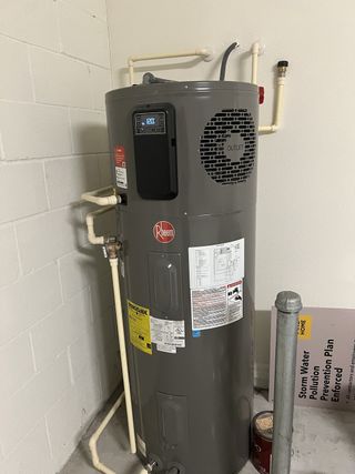 A modern water heater with digital display in a utility room, featuring exposed pipes and a concrete floor.