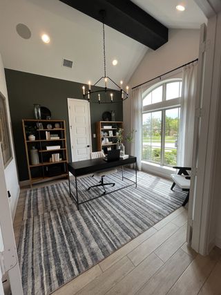 A modern home office with large arched window, dark accent wall, striped rug, and elegant chandelier lighting.