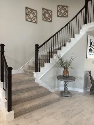 Model Home A modern staircase with elegant railings, framed wall art, and an accent table with a decorative vase on wood-look tile flooring.