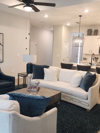 A cozy living room with plush white seating, navy accents, and a rustic coffee table. Open kitchen view with modern lighting.
