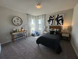 A cozy bedroom with a chic bed, mid-century desk, large clock, abstract art, and warm lighting.