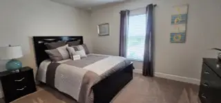 A cozy bedroom with a plush bed, elegant lamp, and soothing wall art, filled with natural light through large windows.