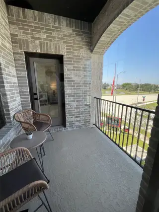 A cozy balcony with wicker chairs and a small table, surrounded by gray brick walls, overlooking a sunny street.
