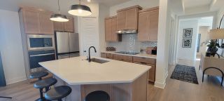 Model Home A modern kitchen with a sleek island, pendant lighting, light wood cabinetry, and a stainless steel fridge.