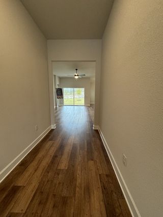 A bright hallway with warm wood flooring leading to a spacious living room with natural light and a ceiling fan.