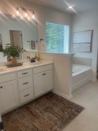 Model Home A bright bathroom featuring a built-in tub, elegant rug, spacious vanity with dual sinks, and chic lighting.