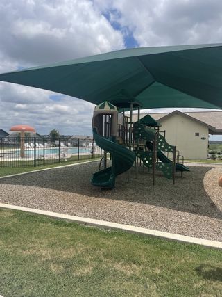 Community Amenities A shaded playground in the Knox Ridge community by Lennar (Converse, TX), featuring green slides, a climbing structure, and a view of the community pool.