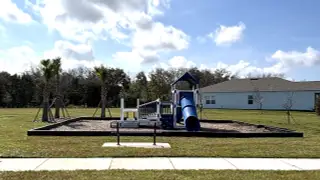 A modern playground with a blue slide and climbing structures, set in a well-maintained green space with palm trees.