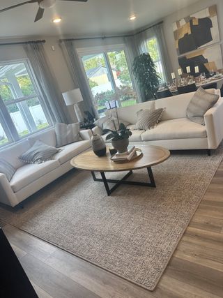 A bright, modern living room with white sofas, a round wooden coffee table, and large windows bringing in natural light.