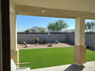 A cozy covered patio with a view of a landscaped yard featuring artificial turf and desert plants.