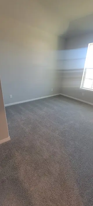 A bright room with plush gray carpet, soft white walls, and ample natural light from a large window.