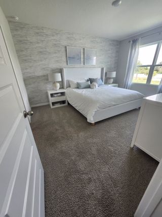 A serene bedroom with a plush carpet, modern white furniture, and soft natural light filtering through large windows.