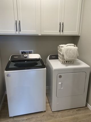 Model Home A modern laundry room features a Whirlpool washer and dryer set, white cabinetry with black handles, a woven basket with towels, and light wood-look flooring.