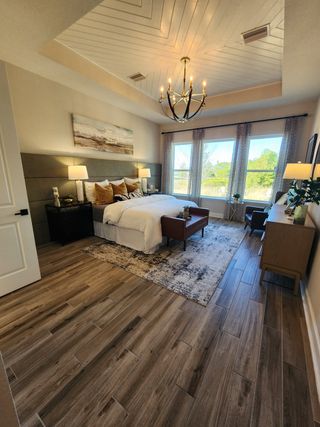A grand primary bedroom with wood flooring, a statement chandelier, and a tray ceiling, radiating modern elegance.