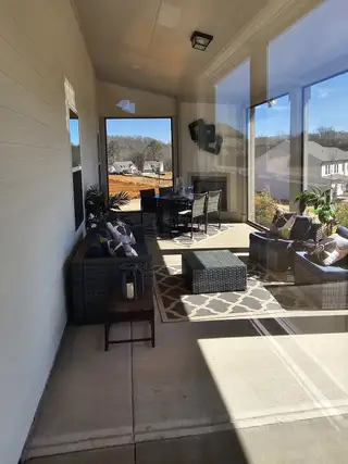 A bright, enclosed patio with large windows overlooking a scenic view, featuring comfortable wicker furniture, a dining set, and a patterned rug.