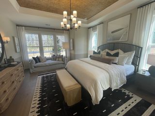 A cozy bedroom featuring elegant bedding, a stylish chandelier, a patterned rug, and ample natural light from large windows.
