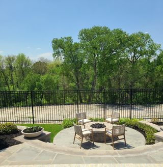 A serene outdoor patio with a fire pit and seating, surrounded by lush greenery and a black iron fence.