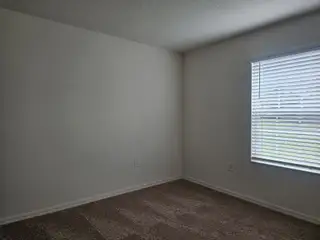 A cozy room with neutral walls, carpeted flooring, and a bright window with blinds filtering soft natural light.