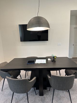 A modern dining area with a sleek black table, gray chairs, a large pendant light, and a wall-mounted TV.