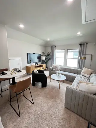 A cozy living space with modern furniture, neutral tones, plush carpet, and natural light filtering through wide windows.
