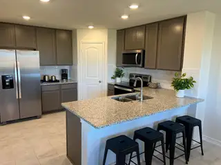 Model Home A modern kitchen in a Medina Crossing home, featuring gray cabinets, granite countertops, stainless steel appliances, a kitchen island with bar stools, and light tile flooring (Von Ormy, TX).