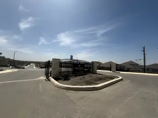 Street view A gated entrance to Magnolia Village (San Antonio, TX) by Rosehaven Homes features a black iron gate with stone pillars under a bright, sunny sky.