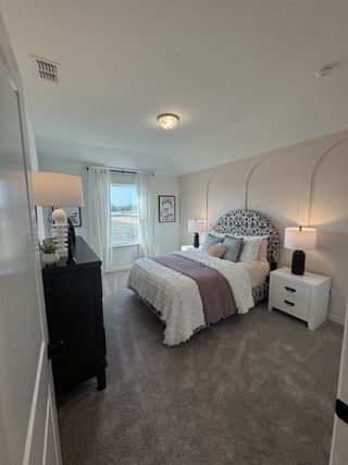 Model Home A cozy bedroom with a patterned headboard, plush bedding, and soft carpeting, accented by natural light from a large window.