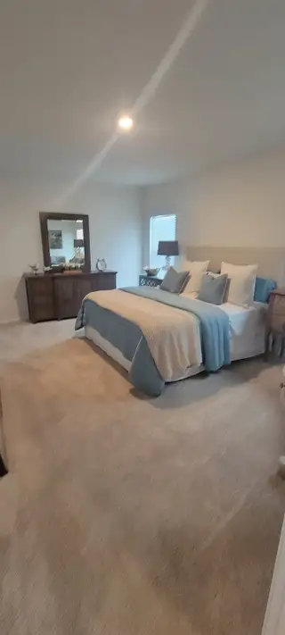 A spacious bedroom with neutral carpet, a large bed with soft blue accents, and a dark wood dresser creates a comfortable retreat.
