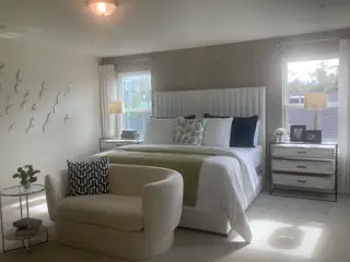 A chic bedroom featuring a plush headboard, elegant wall art, and cozy seating with natural light through large windows.