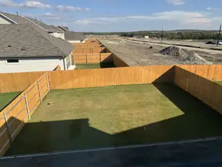 A fenced backyard with lush green grass and a view of the developing neighborhood in Longview by Taylor Morrison (Del Valle, TX).