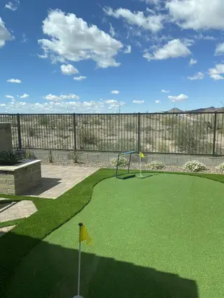 A private backyard oasis features a putting green with flags, a paved patio with a fire pit, and a scenic desert view beyond a black metal fence in The Villas at Mystic by Brightland Homes (Peoria, AZ).