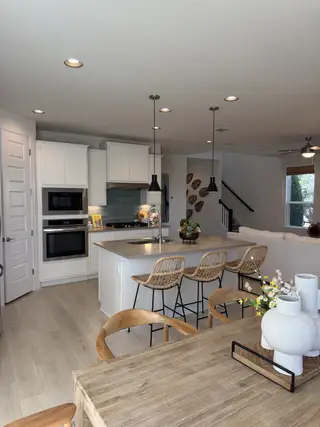 Model Home A modern kitchen with sleek white cabinets, an island, and pendant lighting, flowing into an open dining area with natural wood accents.