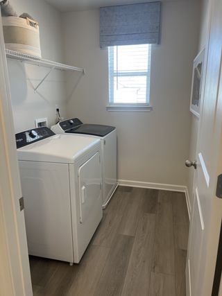 A bright laundry room with a white Whirlpool top-load dryer, matching washer, wire shelving, a woven basket, and a window with a blue shade.