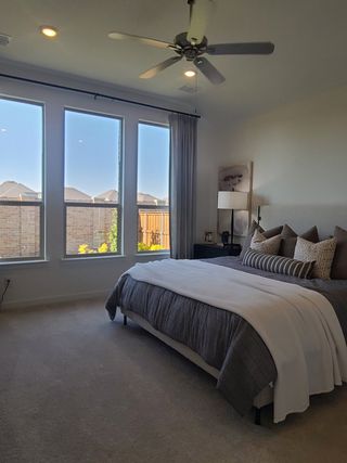 Model Home A cozy bedroom with a large window, neutral bedding, soft carpeting, and a ceiling fan for a relaxing ambiance.