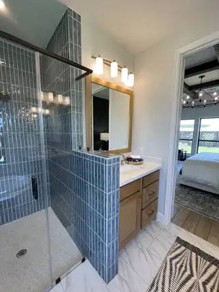 Model Home A modern bathroom with blue-tiled rain shower, wood vanity, elegant lighting, and marble-look flooring.