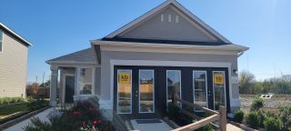 A bright, sunny view of a model home in Mission del Lago by KB Home (San Antonio, TX), featuring a light gray exterior, a paved walkway, and "Welcome" signs in the windows.