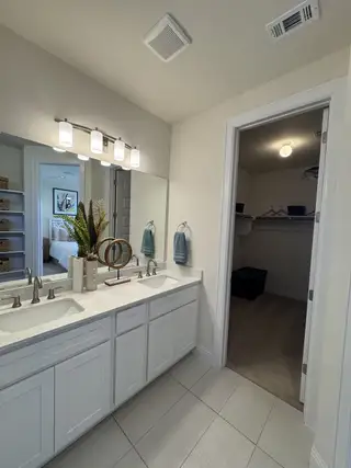 A modern bathroom with double sinks, sleek cabinetry, and soft lighting leading into a spacious walk-in closet.